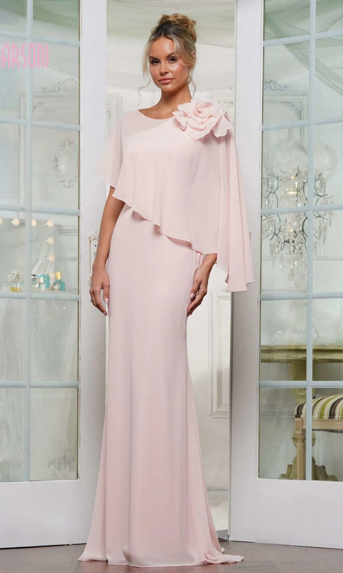 A woman stands indoors in front of glass doors wearing the Marsoni MV1347 Long Formal Dress in blush, featuring floor-length chiffon, an asymmetrical cape, and a large fabric flower on one shoulder.